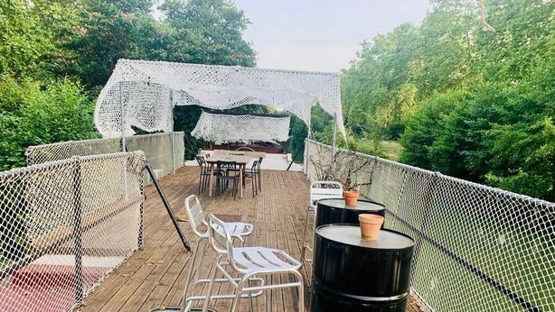 Una terraza exterior con piso de madera, red de malla blanca, sillas de metal, una mesa y un barril negro grande.