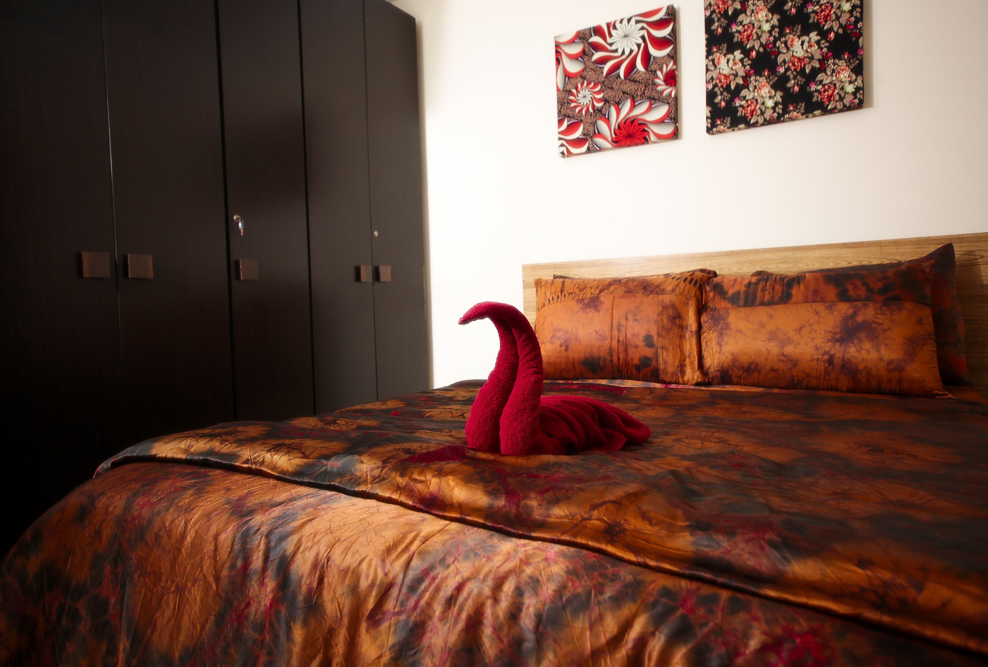 Un lit avec une serviette rouge en forme de cygne, une tête de lit en bois et deux œuvres d’art murales aux motifs floraux.