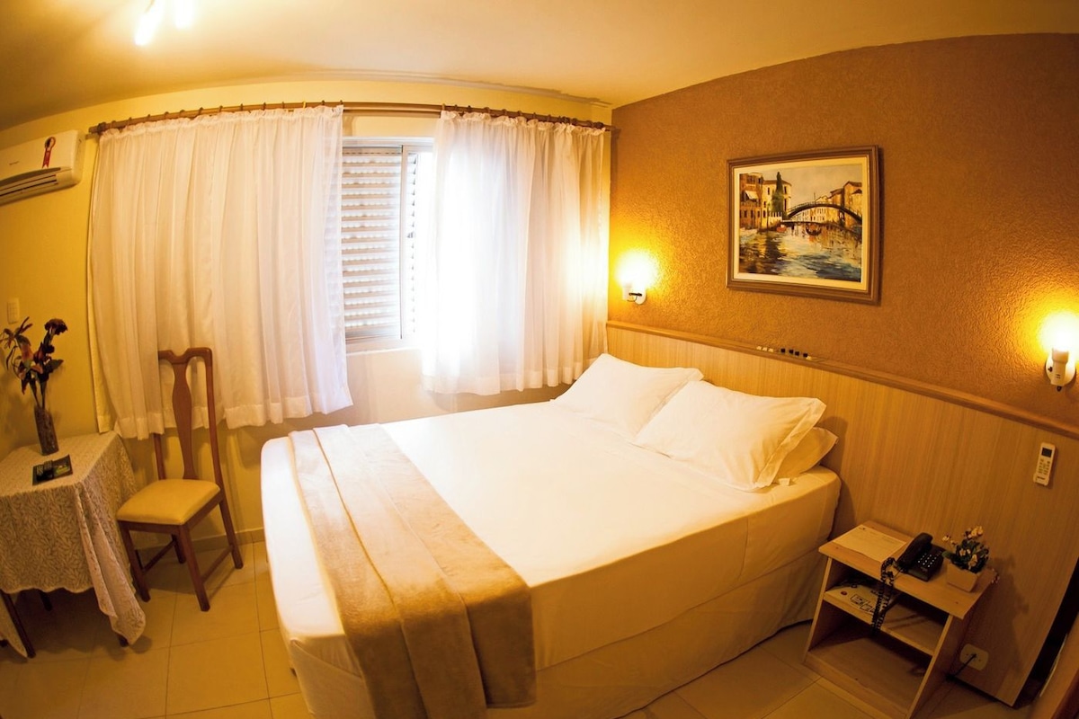 Une chambre d’hôtel avec un grand lit, une table de chevet avec une lampe, une chaise, une petite table avec un vase et un tableau au mur.