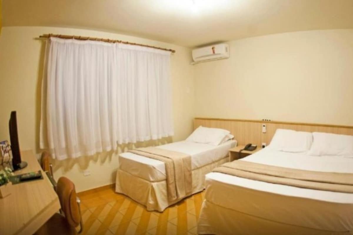 Une chambre d’hôtel avec deux lits, un bureau avec un ordinateur, une chaise, une fenêtre avec des rideaux et un appareil de climatisation.