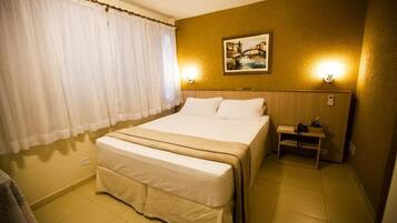 Een hotelkamer met een groot bed, een klein nachtkastje, een aan de muur hangend schilderij en een raam met gordijnen.