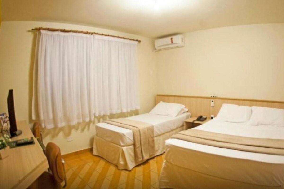 Habitación de hotel con dos camas, un escritorio con computadora, una silla, una ventana con cortinas e un equipo de aire acondicionado.