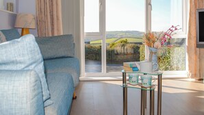 A living room with a blue sofa, a glass coffee table with two mugs, a vase with flowers, and a view of a green landscape through a sliding glass door. - Thornberry (Salcombe)