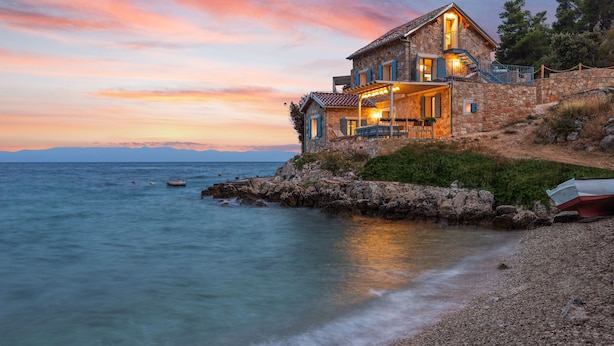 Ein Steinhaus mit einem Balkon, der Blick auf eine felsige Küstenlinie und ruhiges Meer bietet.