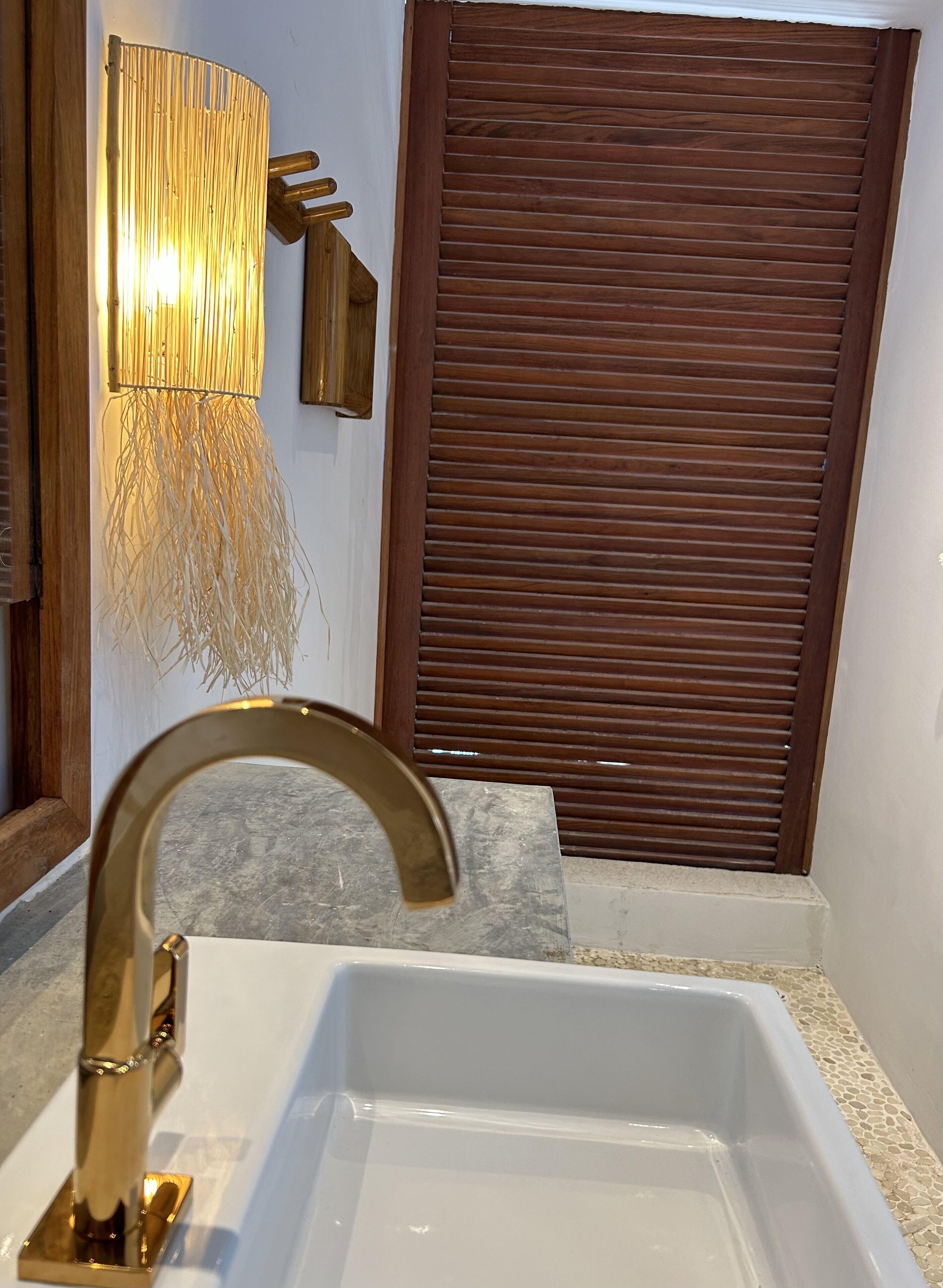 Un baño con lavamanos blanco, grifo dorado y aplique de pared con diseño de bambú.