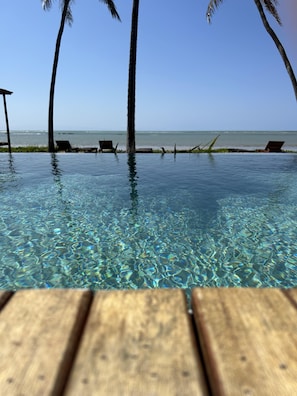 A clear pool with wooden deck in the foreground, palm trees, and a beach in the background. - Kuarasy Boutique Hotel Japaratinga (Japaratinga)