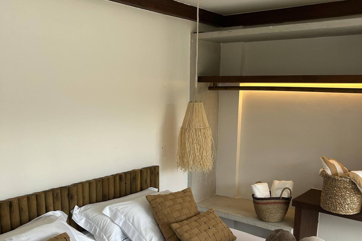Une chambre d’hôtel avec un lit, des oreillers, un luminaire suspendu en fibres tressées et une petite table sur laquelle se trouvent un panier et des serviettes.