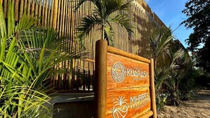 Edessä on bambuaidasta ja palmuista muodostuva maisema, ja sen edessä on puinen kyltti, jossa on logot majoitusliikkeelle »Pousada Kuarasy» ja rantaklubille »Mhares Beach Club».