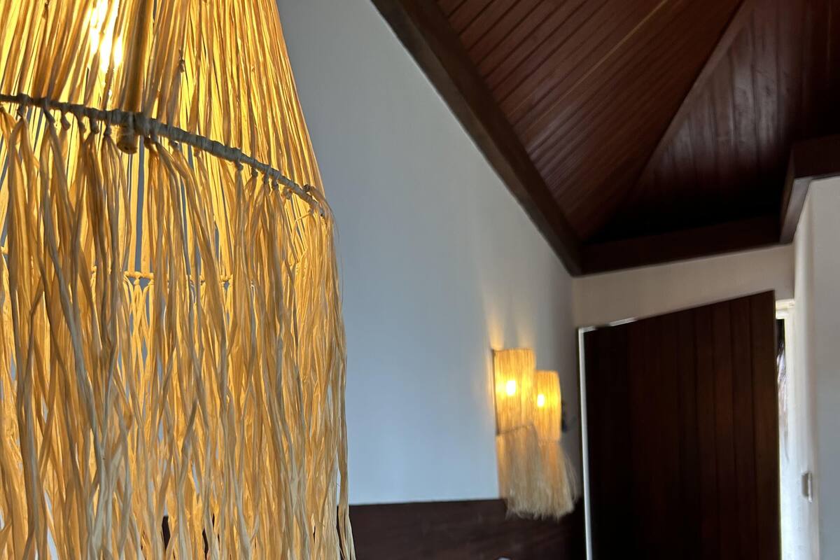 Une chambre à coucher avec un lit, du linge de lit blanc, une tête de lit en osier et un luminaire suspendu.