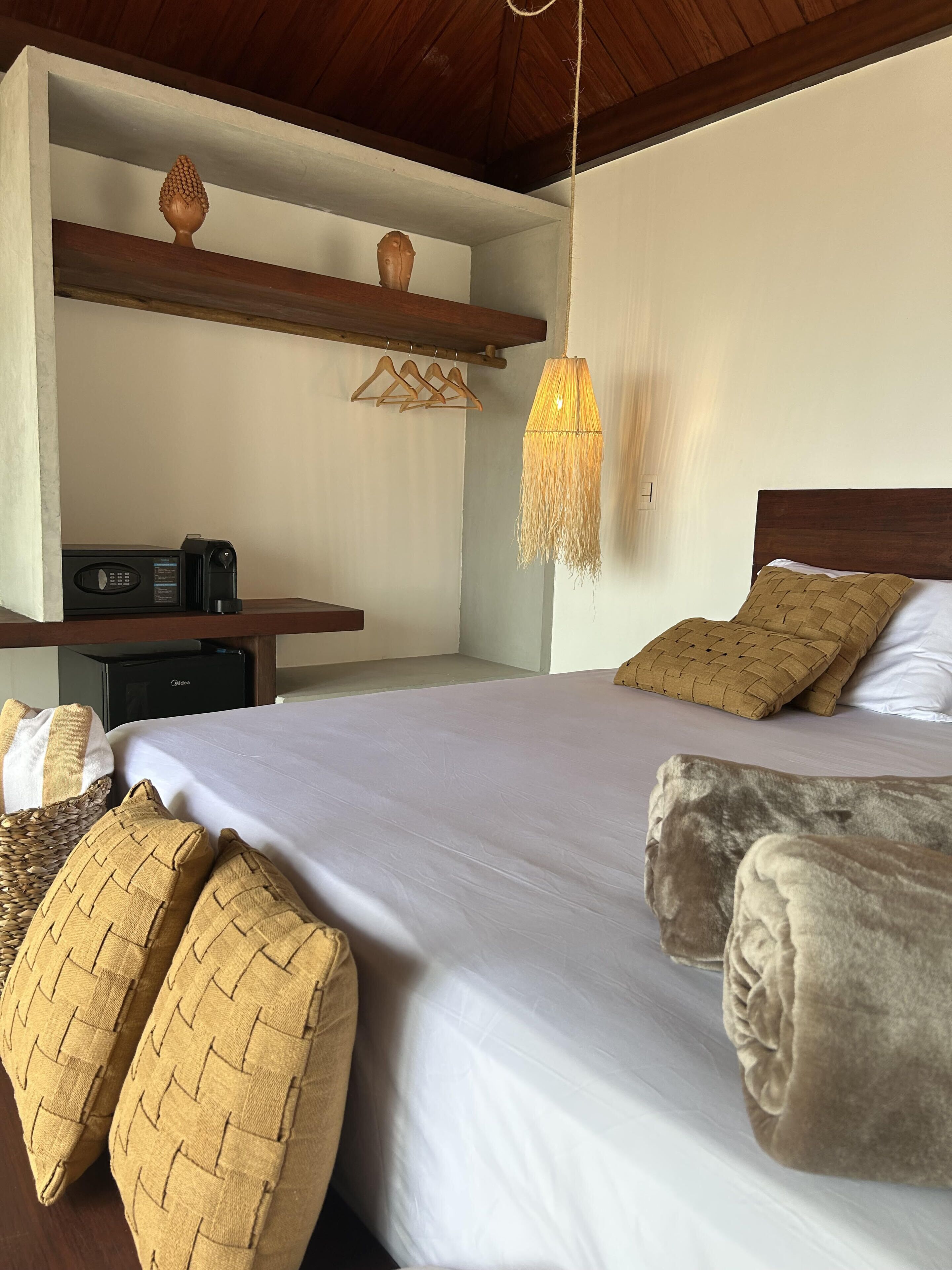 Un dormitorio con una cama grande, cabecera de madera, lámpara colgante, estante con adornos y mesita de noche con radio y cafetera.