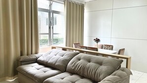 Ein modernes Wohnzimmer mit einem beigefarbenen Ecksofa, einem Esstisch mit Stühlen und einem großen Fenster mit Vorhängen.