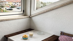 Una esquina de una habitación con una ventana, una mesita con un vaso de leche, un biberón y una silla con un cojín estampado.