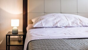 Una cama bien hecha con sábanas blancas y un cubrecama acolchado, una lámpara de noche sobre una mesita y un cabecero con acabado de madera oscura.
