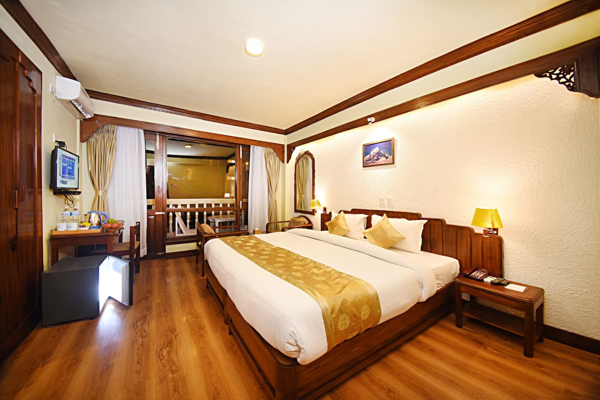 Une chambre d’hôtel avec un grand lit, un plancher en bois, un bureau avec un ordinateur, une chaise, un petit réfrigérateur et une porte donnant sur un balcon.