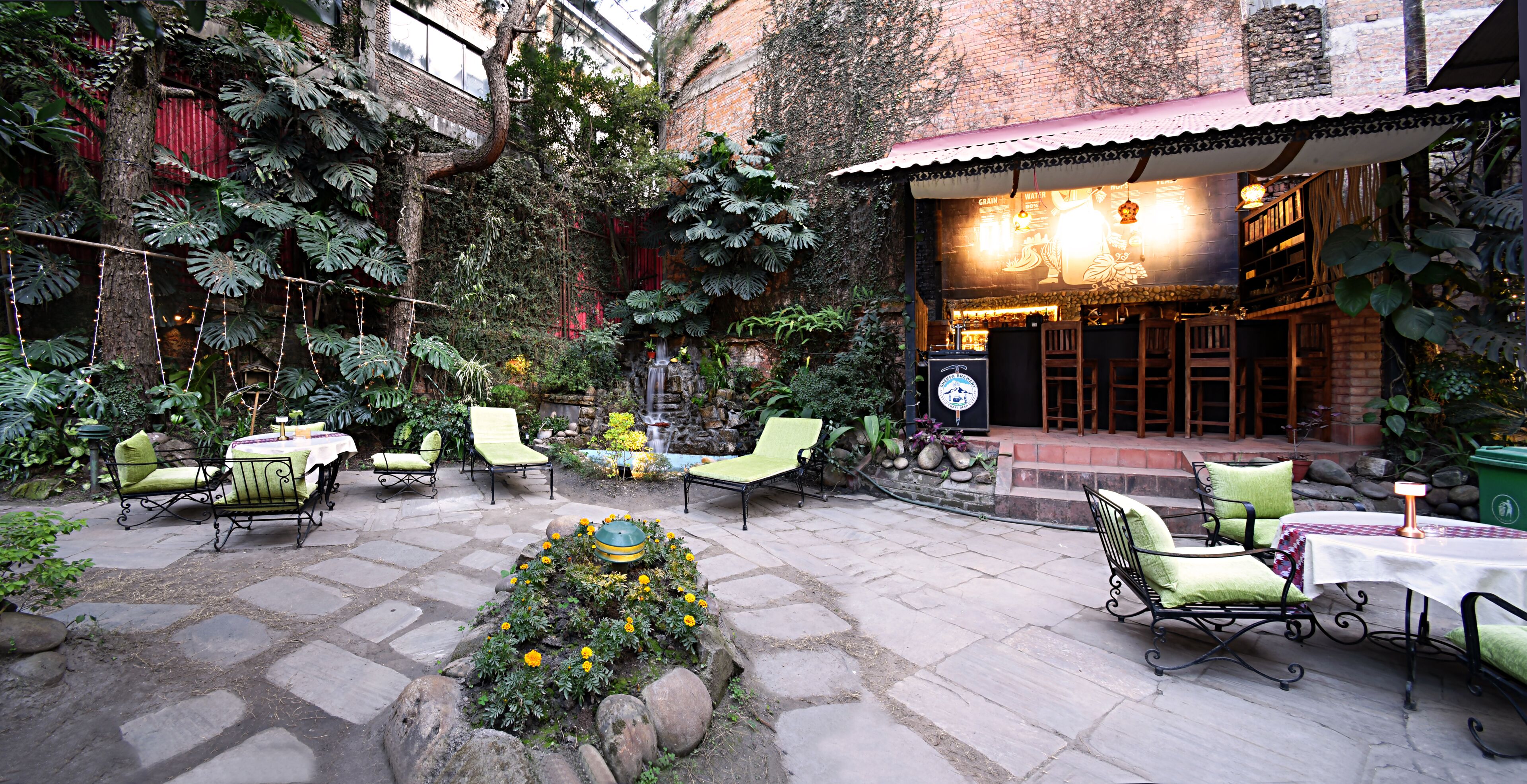 An outdoor patio with stone flooring, green cushioned chairs, a small fountain, and a restaurant with a red roof.