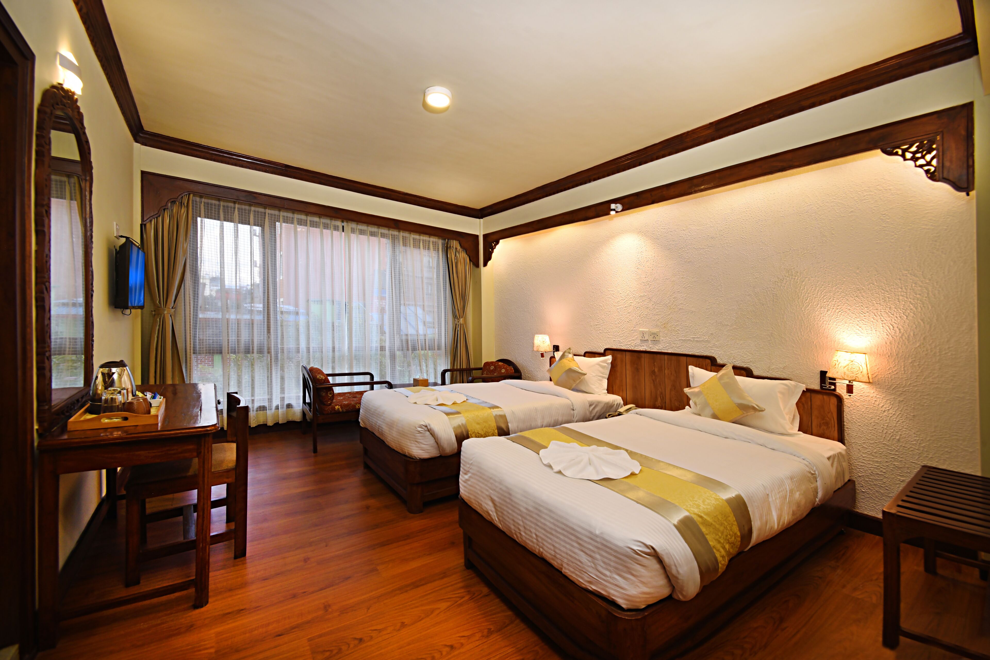 A hotel room with two beds, a wooden desk, a chair, a table with a lamp, and a window with curtains.