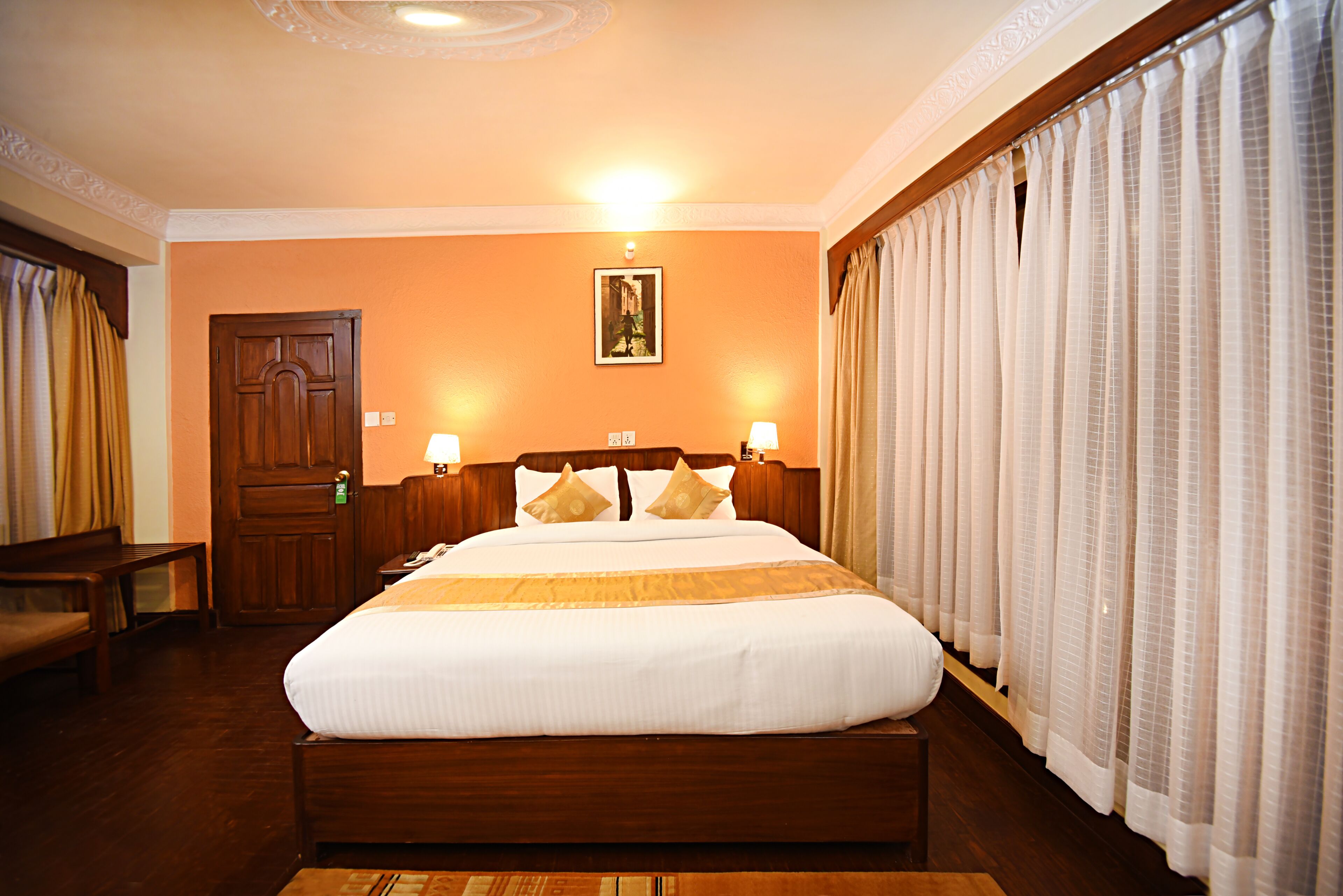 Une chambre d’hôtel avec un grand lit, une tête de lit en bois, deux lampes de chevet, un banc en bois et une porte.