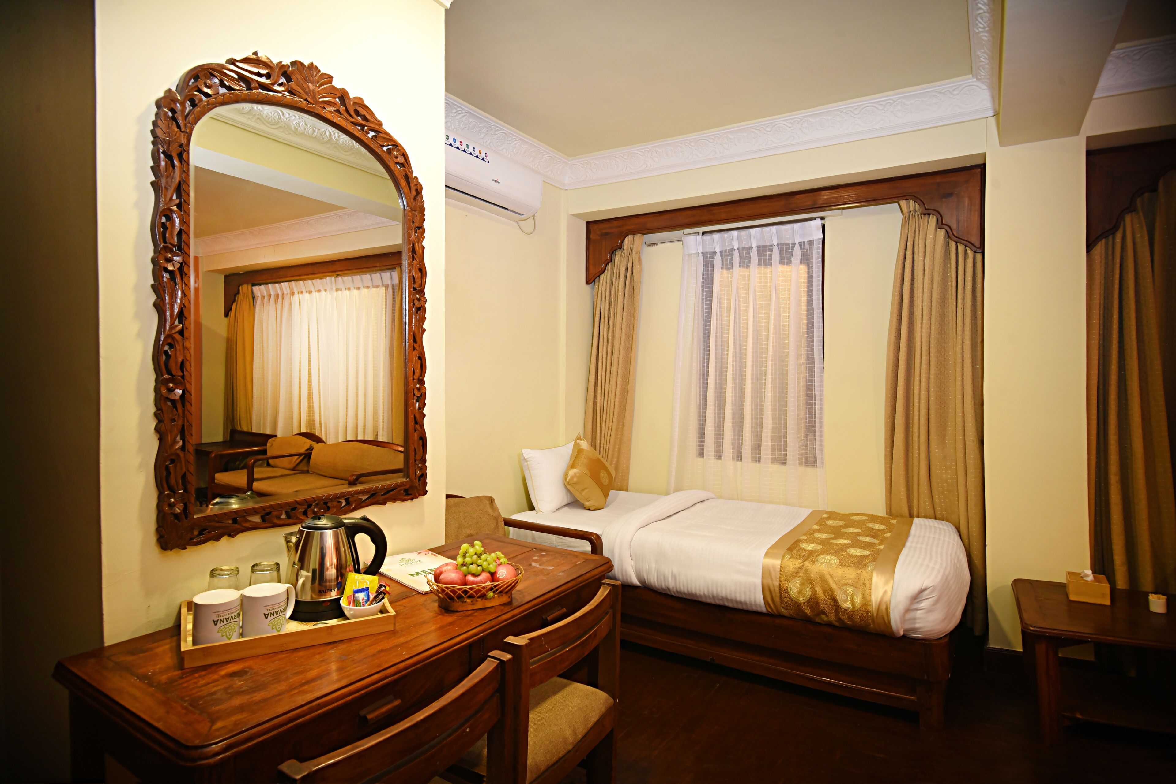 A hotel room with a wooden desk, a bed with a gold blanket, a large mirror, and a window with curtains.