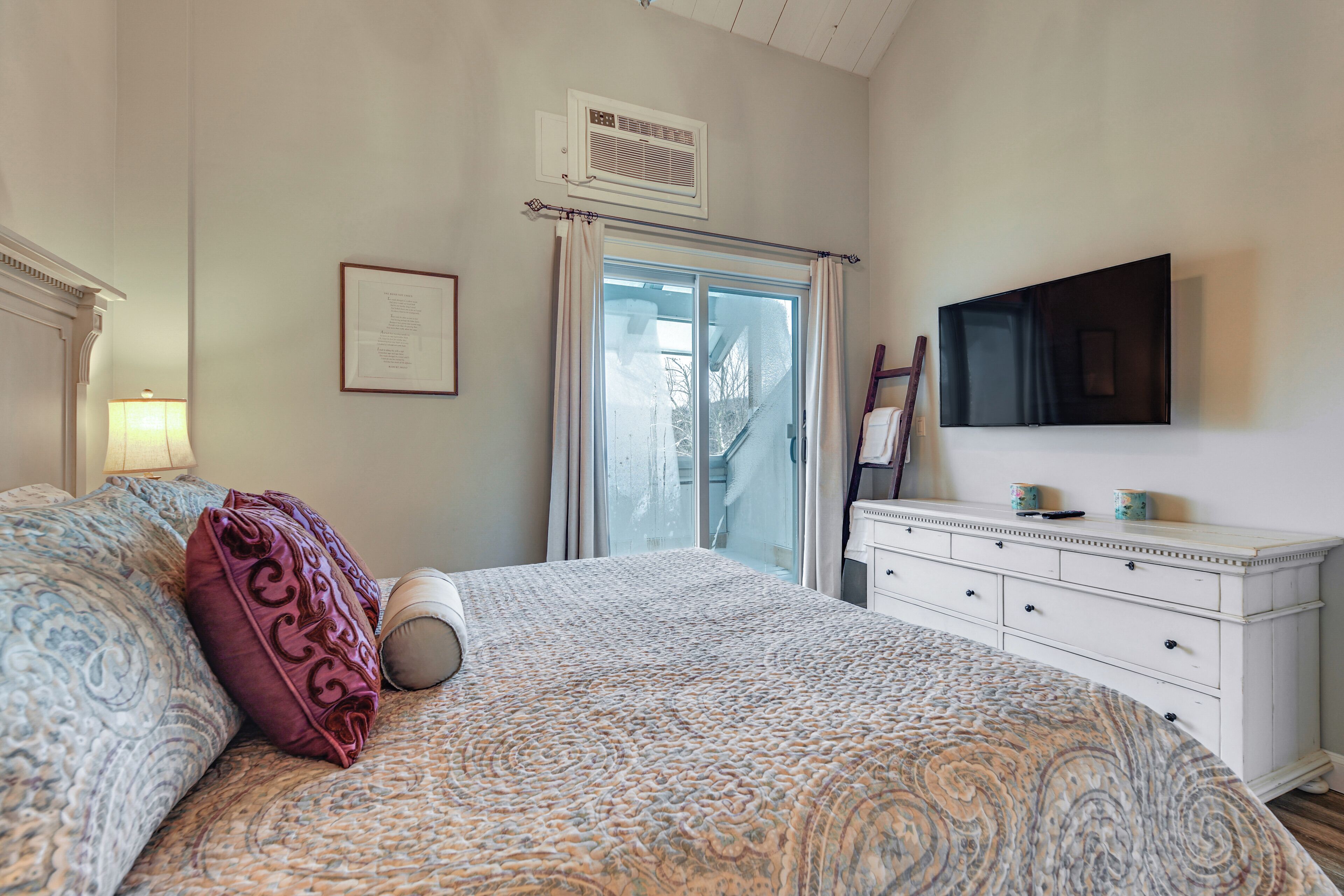 A bedroom with a large bed, a dresser, a flat-screen TV, and a window with curtains.