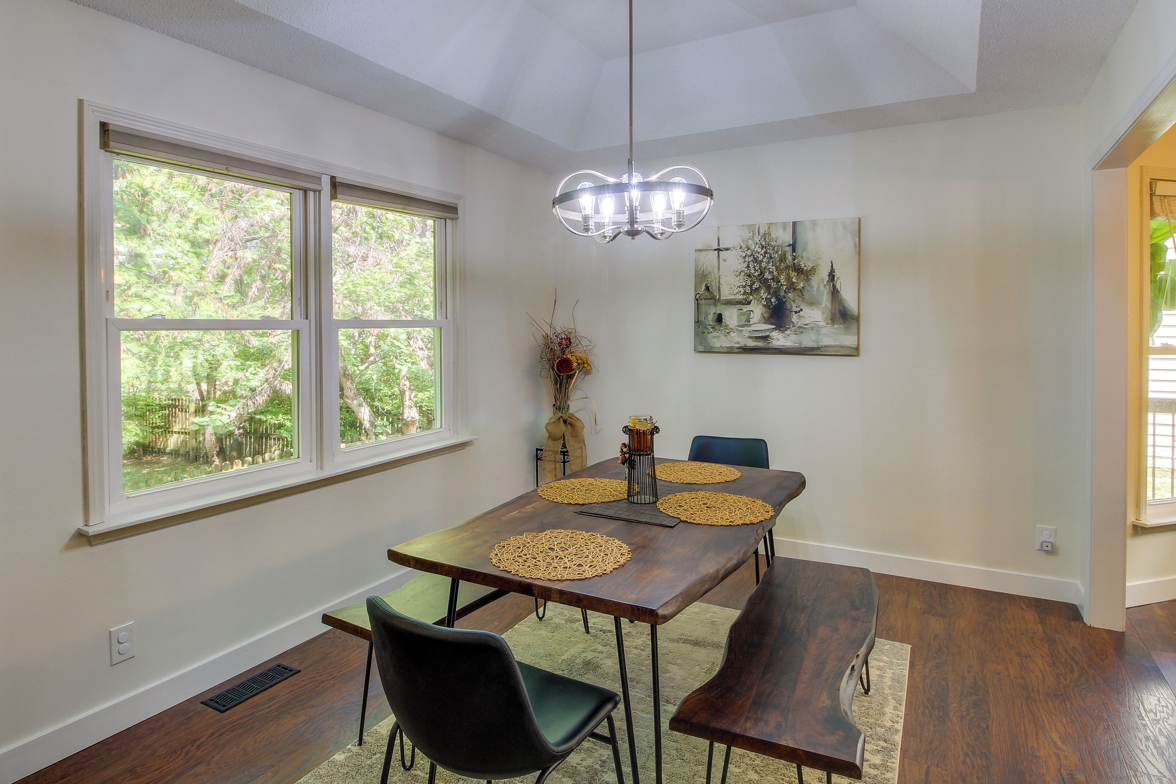 A dining area with a wooden table, four chairs, a chandelier, and a painting on the wall.