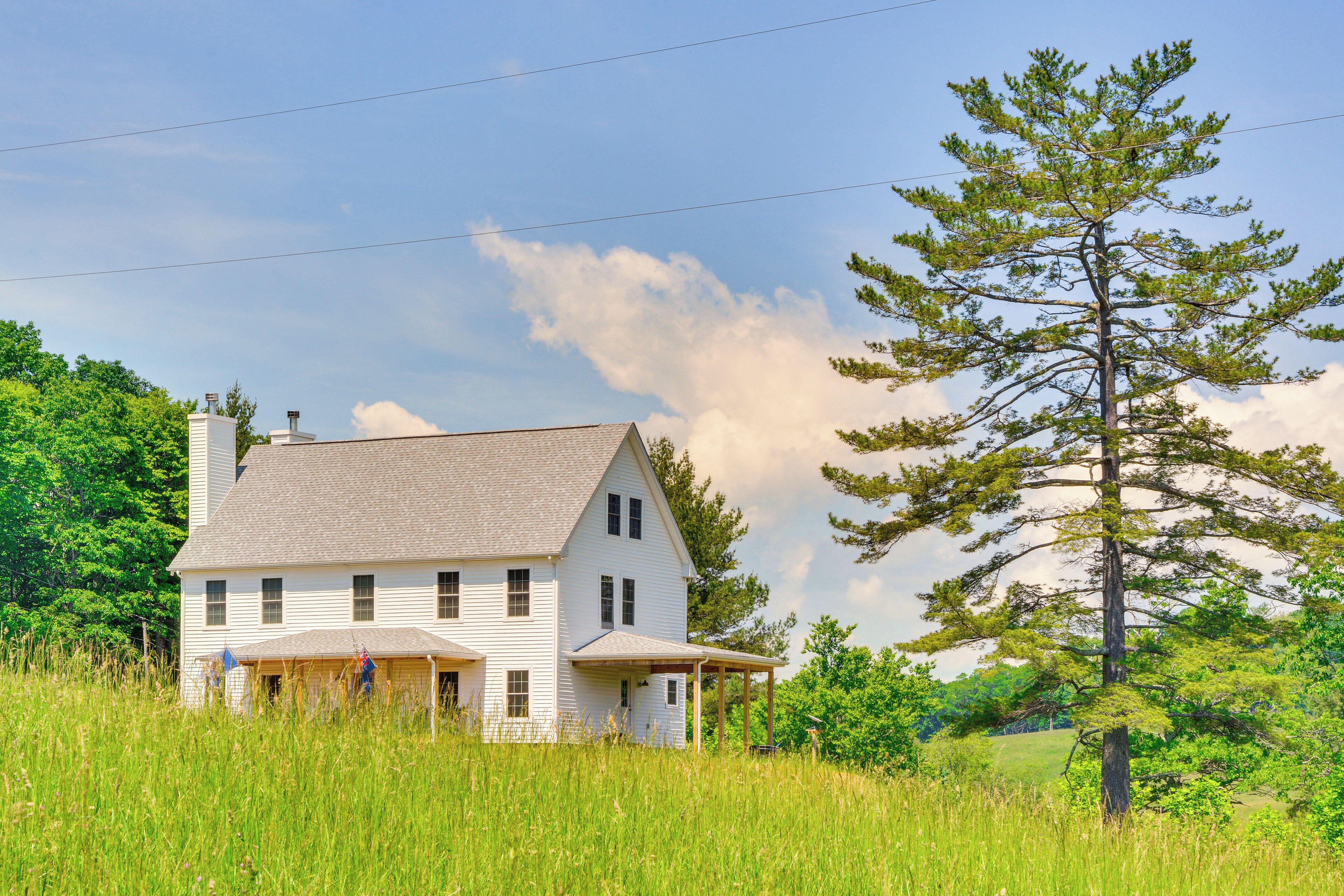A white two-story house with a gabled roof, a covered porch, and a large pine tree in the foreground.