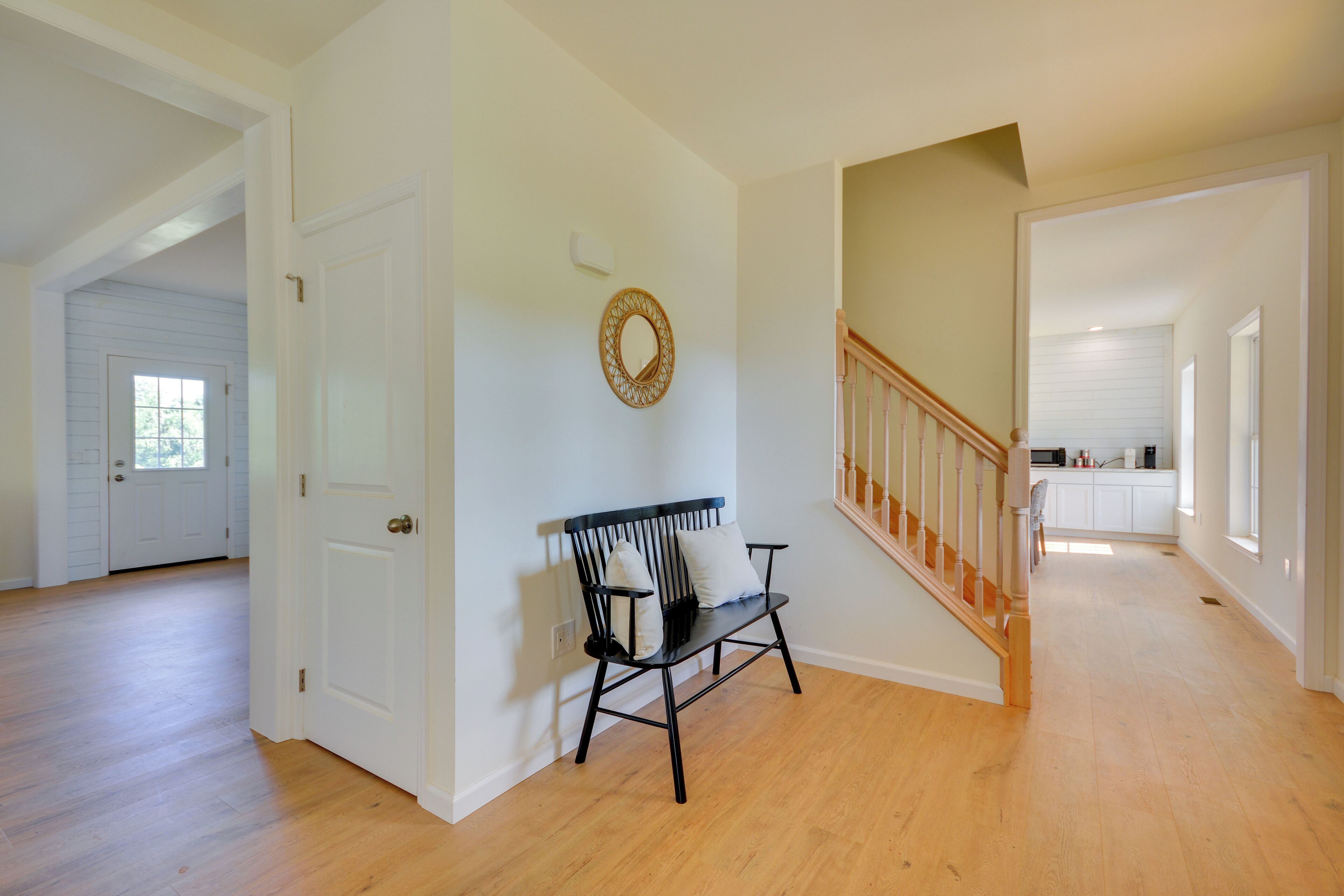 A spacious hallway with hardwood floors, a black bench with cushions, a decorative mirror, and a staircase leading to a kitchen area.