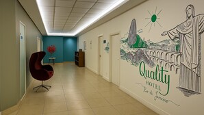 A hotel hallway with a red chair, a small table with a vase, and a mural on the wall. - Flat Próximo ao Riocentro - Barra da Tijuca (Rio de Janeiro)