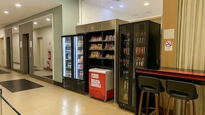 A hotel lobby with a self-service beverage station, a refrigerator stocked with snacks, and a bar area with stools. - Flat Próximo ao Riocentro - Barra da Tijuca (Rio de Janeiro)