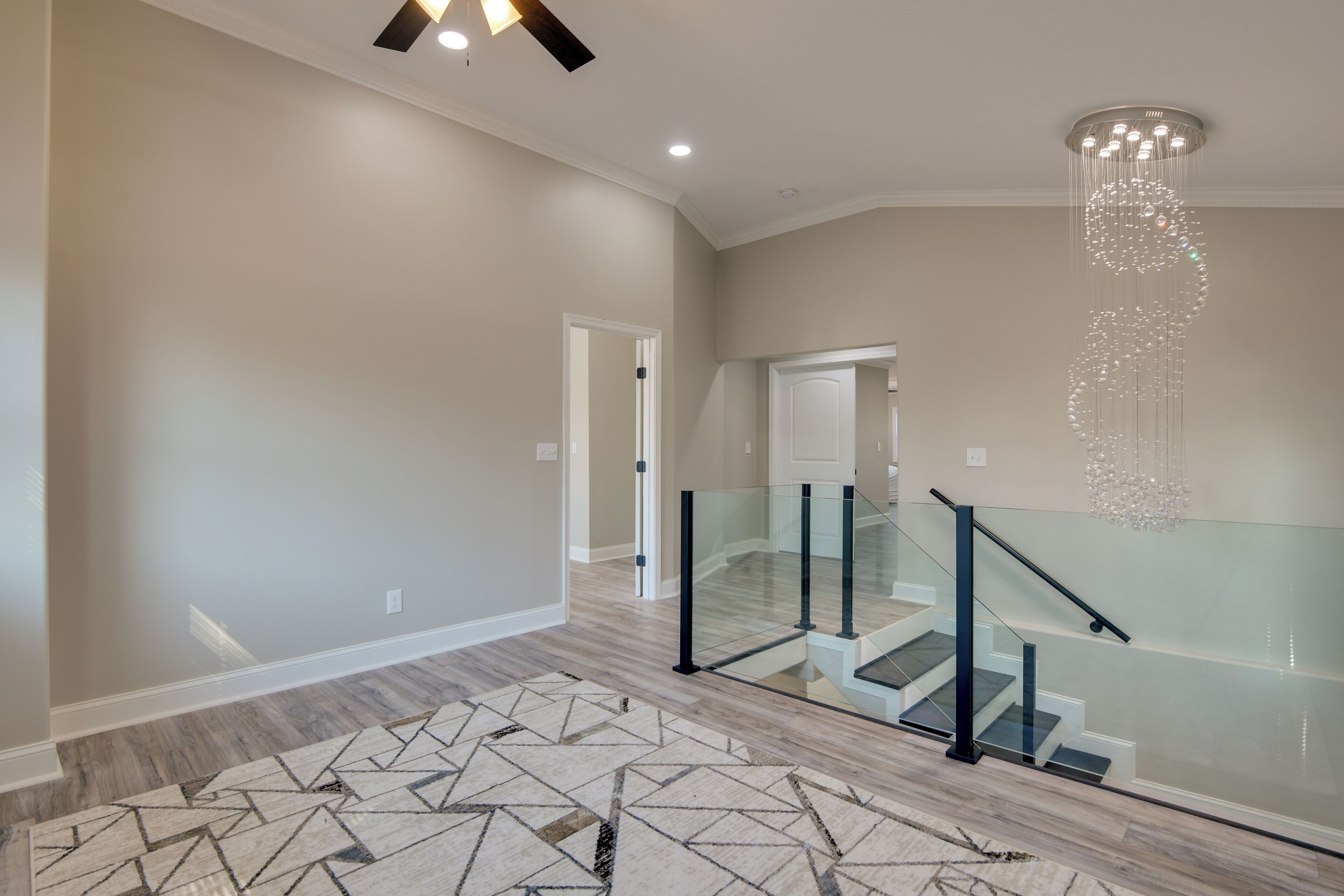 A spacious hallway with a geometric patterned rug, a modern staircase with glass railings, and a decorative chandelier.