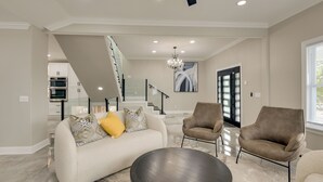 A spacious living room with a sofa, two armchairs, a coffee table, and a chandelier. A staircase leads to an upper level with a kitchen and a dining area. - 5 Mi to Inner Harbor: Expansive Family Getaway (Baltimore)