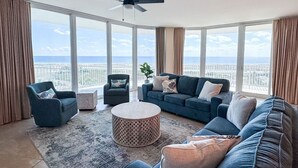 A spacious living room with a large window offering a city view, a ceiling fan, and a cozy seating arrangement. - DOWN BY THE BAY! 4 Bedroom Gulf Facing unit! ASK ABOUT DISCOUNTS!  (Orange Beach)