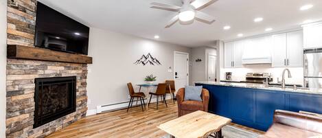 A modern living room with a stone fireplace, wooden flooring, a dining area, and a kitchen with blue cabinets.