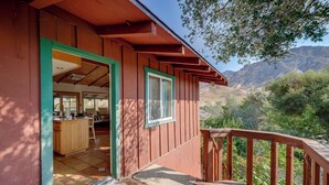 A wooden cabin with a porch, a kitchen area visible through an open door, and a view of mountains in the background. - Scenic Kernville Getaway w/ Deck & Mountain Views! (Kernville)