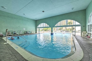 An indoor swimming pool with clear water, a tiled floor, and a view of the outside through large arched windows.