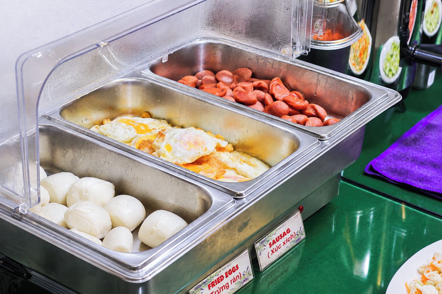 A buffet with labeled food sections including fried eggs, sausages, and dumplings.