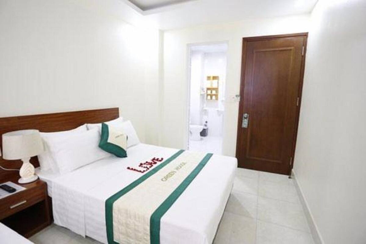 A hotel room with a neatly made bed, a wooden headboard, a bedside table with a lamp, and a door leading to a bathroom.