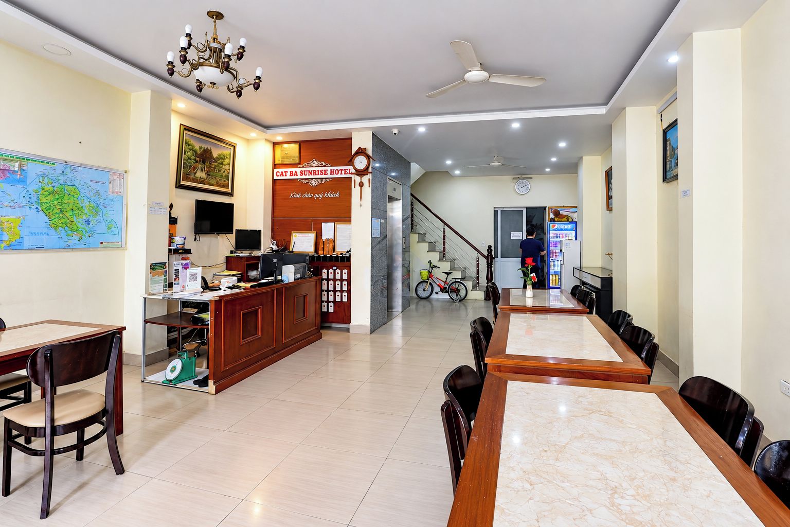 A hotel lobby with a reception desk, a dining area with a large table and chairs, a staircase, and a bicycle.