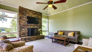 A living room with a stone fireplace, a ceiling fan, a large window, and a couch with pillows. - Lake-view Home at Margaritaville Resort! (Osage Beach)