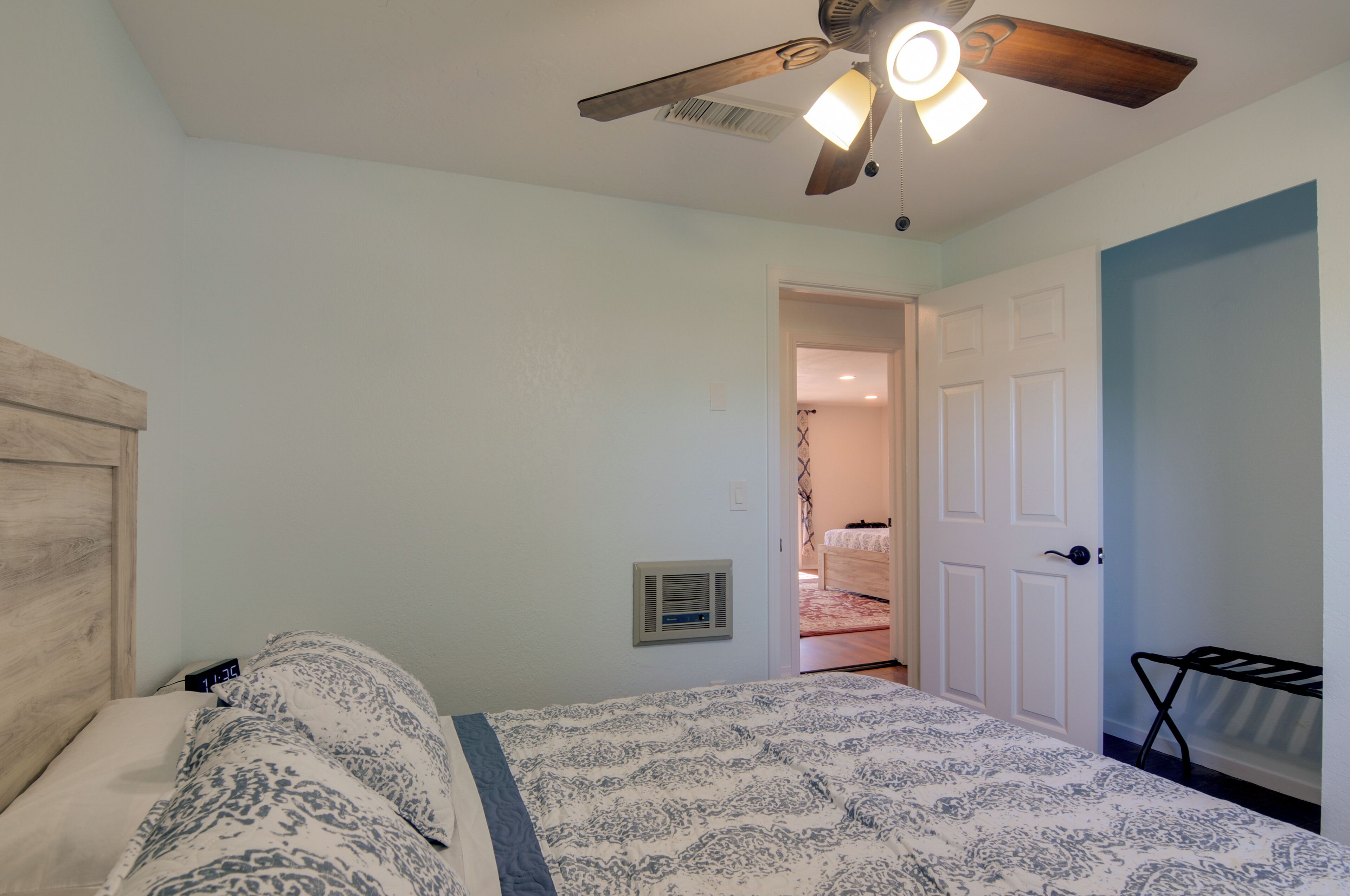 A bedroom with a bed, a ceiling fan, a wall-mounted heater, and a door leading to another room.