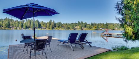 A deck with a table and chairs, an umbrella, and a view of a lake and forest.