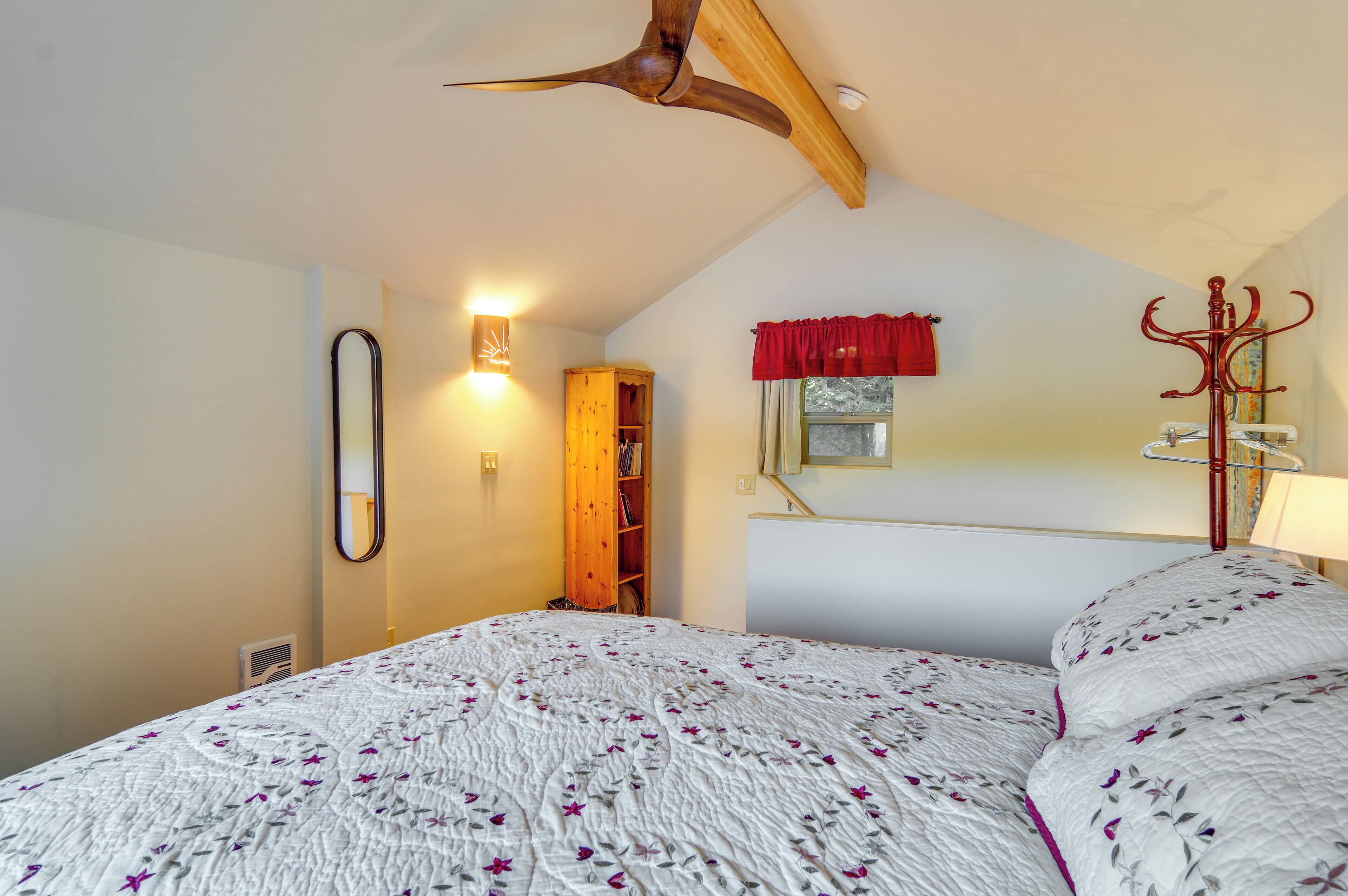 Un dormitorio con una cama, un ventilador de techo de madera, un espejo, una cortina roja y un perchero.