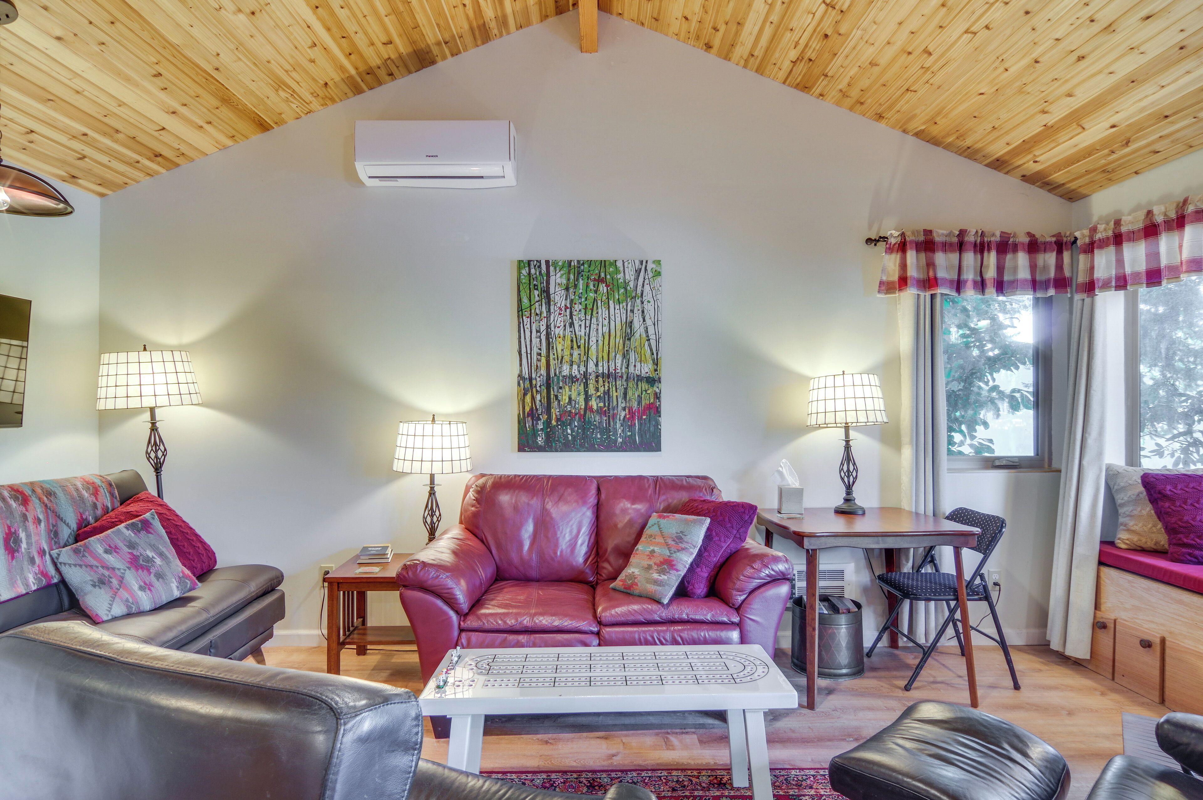 A cozy living room with a red leather sofa, a white coffee table, and a dining table with chairs. There is a painting on the wall and a window with curtains.