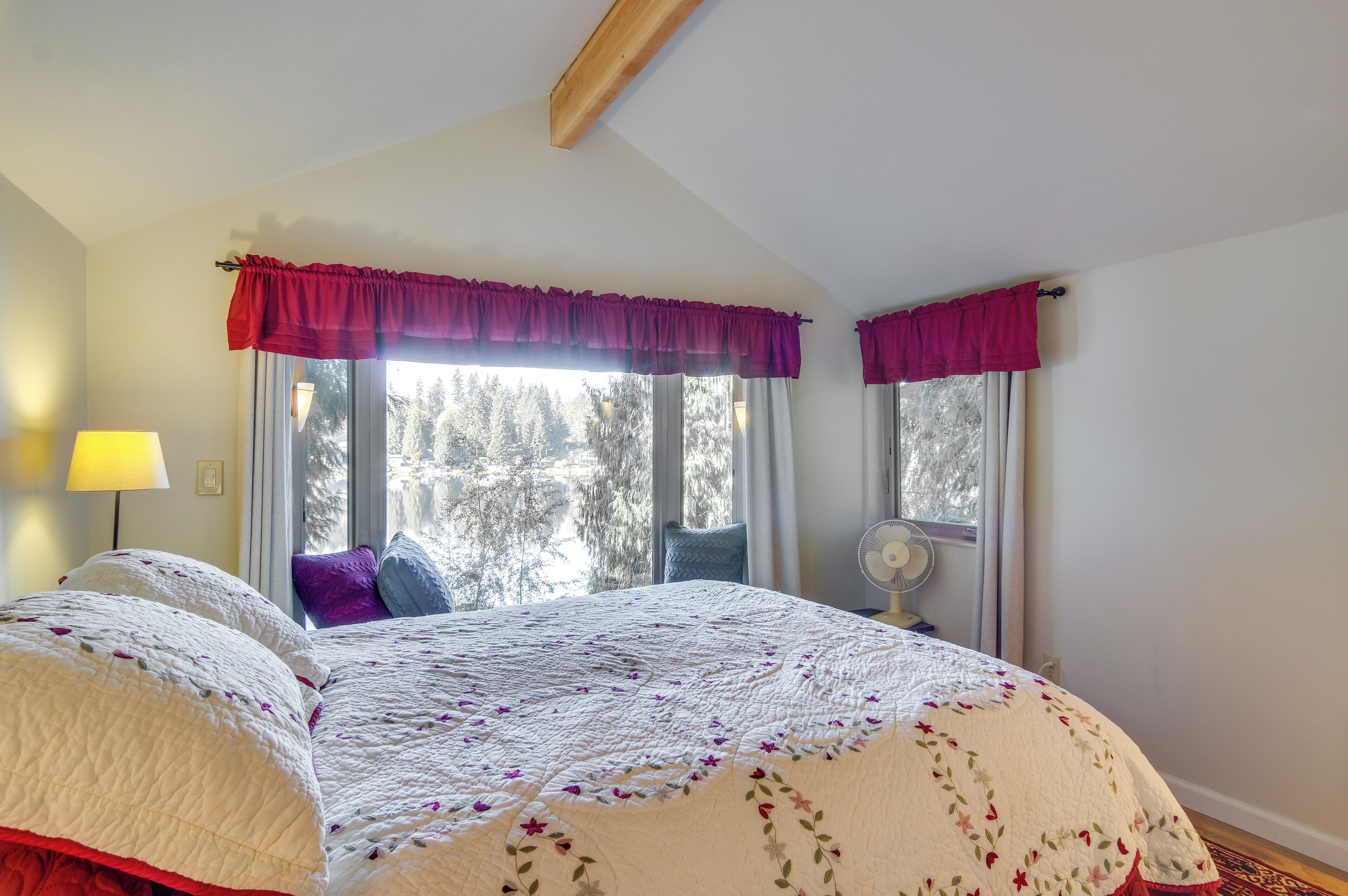 A bedroom with a large bed, a window with curtains, a lamp, and a fan.