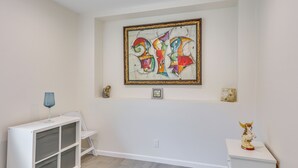 A room with a white wall, a colorful abstract painting, a small statue, and a blue glass on a white cabinet. - Cozy Escondido Escape Near Hiking & Downtown! (Escondido)