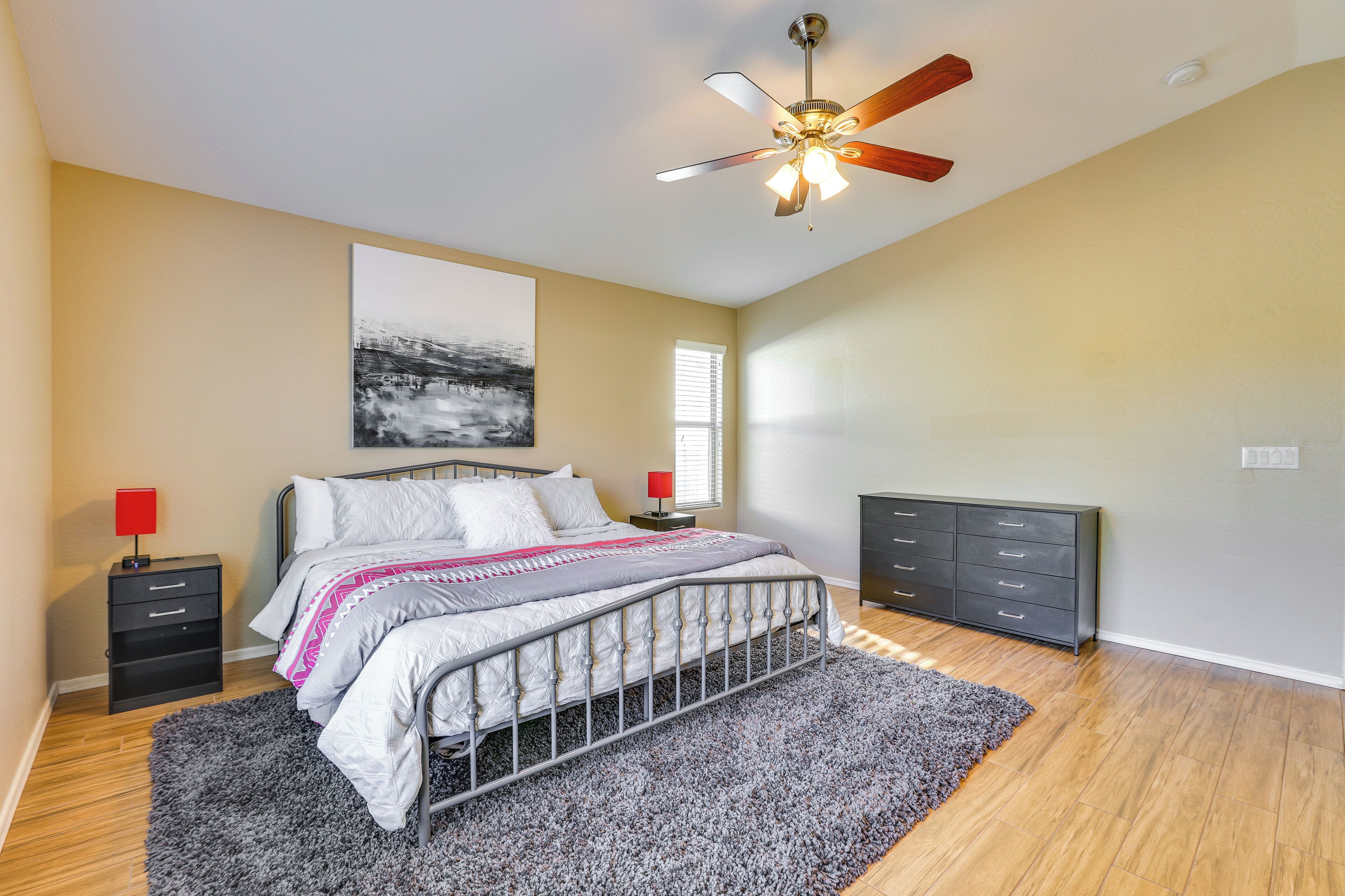 Une chambre à coucher comprenant un lit, des tables de chevet, une commode, un ventilateur de plafond et une fenêtre avec des stores.