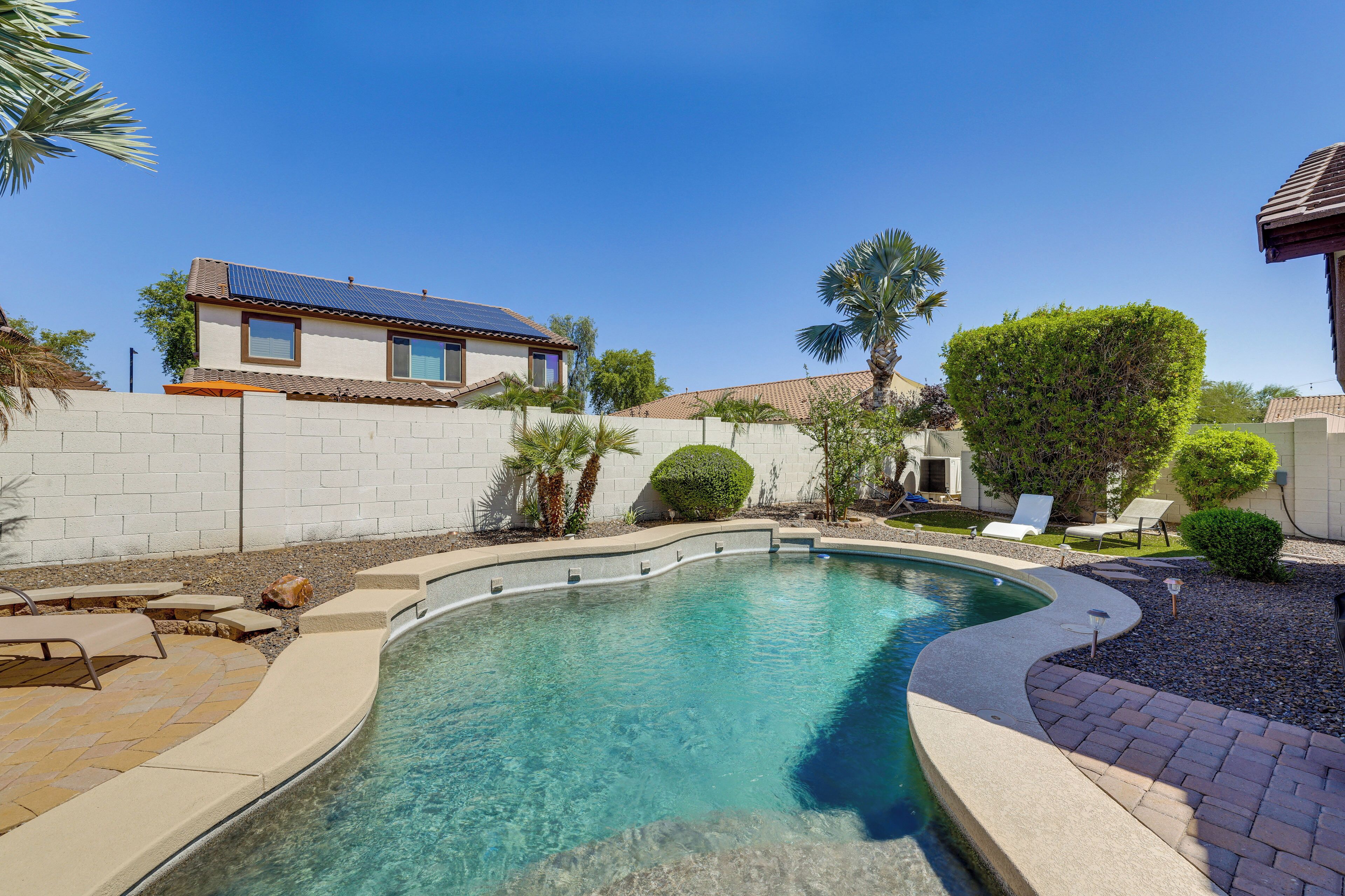 A swimming pool with a curved edge, surrounded by a brick patio, palm trees, and a house with a tiled roof.