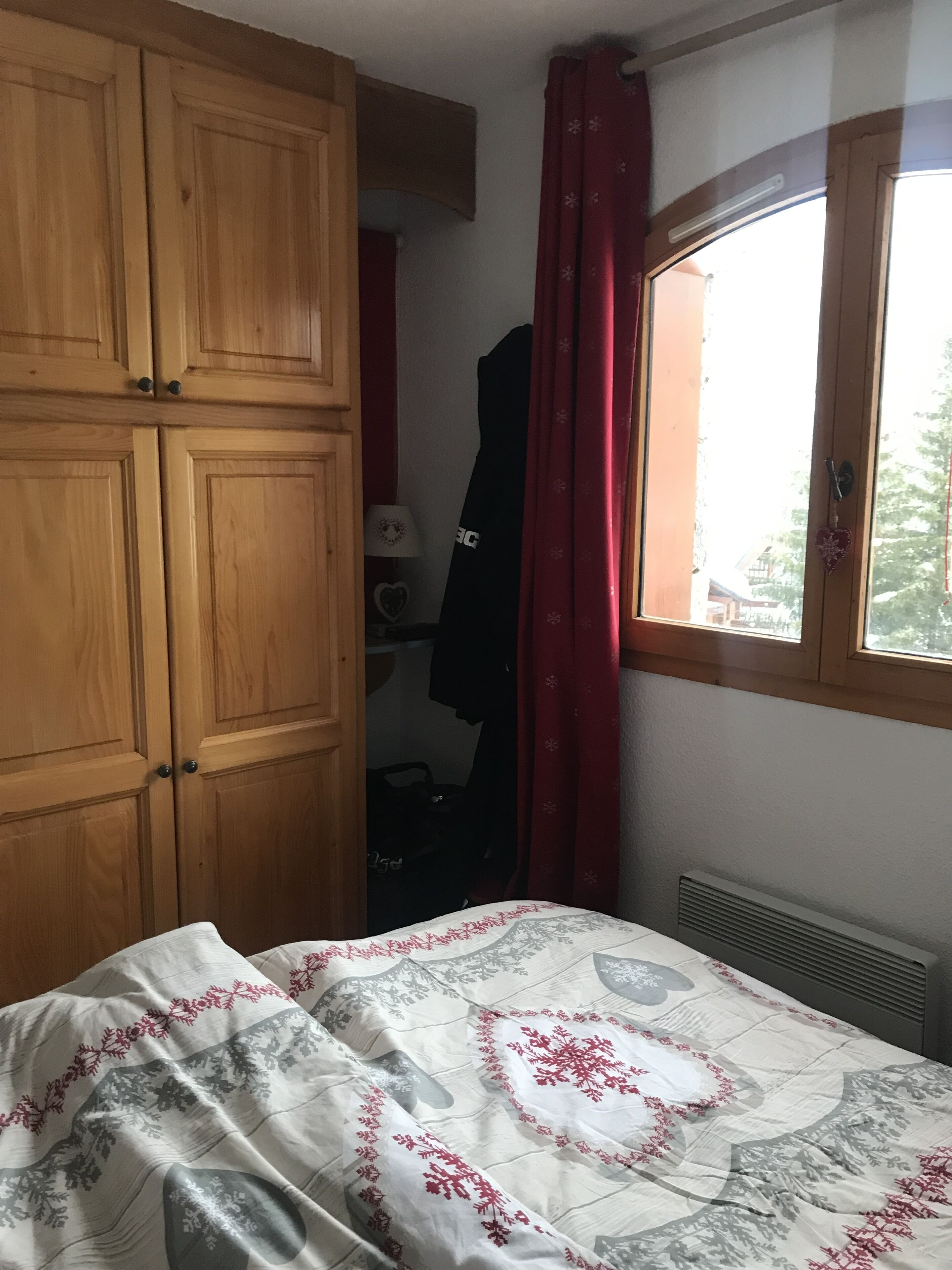 A bedroom with a bed, wooden wardrobe, and a window with red curtains.