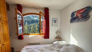 A bedroom with a bed, a window with curtains, a wall-mounted heater, and a painting.