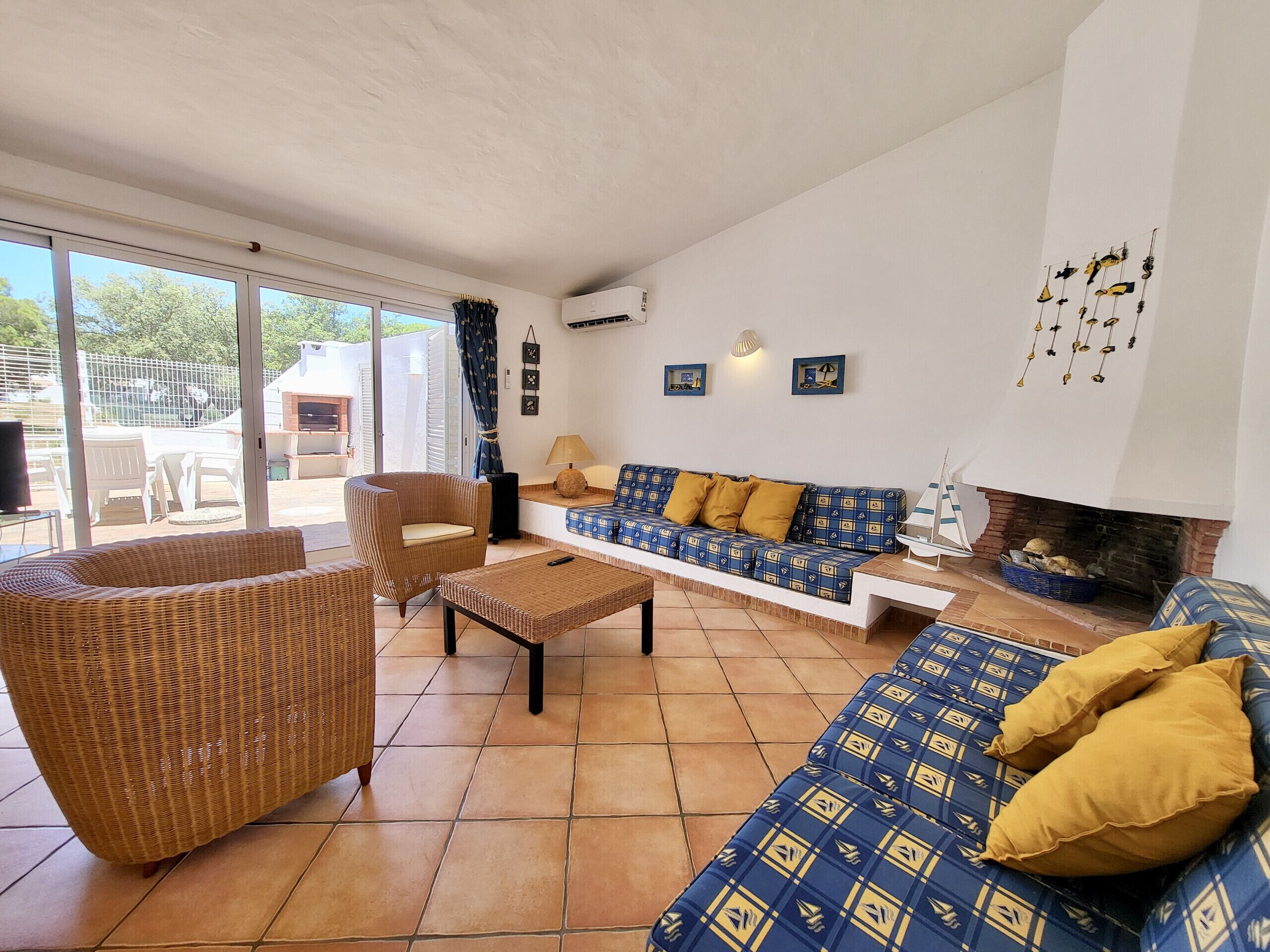 A spacious living room with a large sofa set, a wicker armchair, and a coffee table. There is a fireplace and a view of the outside through large windows.