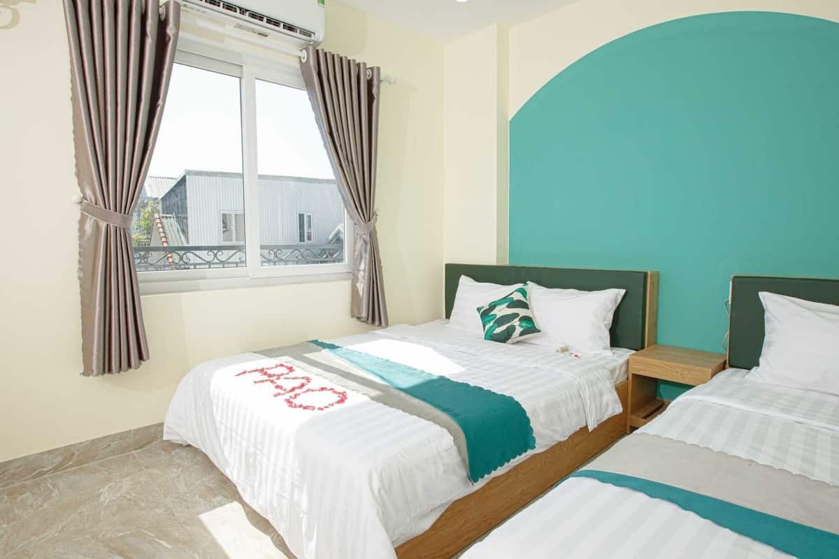 A hotel room with two beds, a window with curtains, a teal accent wall, and a view of buildings outside.